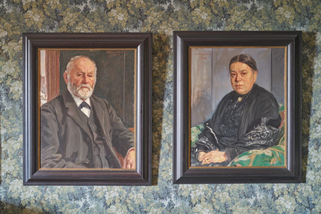 gemalte Porträts von Albert und Bertha Bühler-Reichling als ältere Leute hängen nebeneinander an einer Wand mit Blumentapete